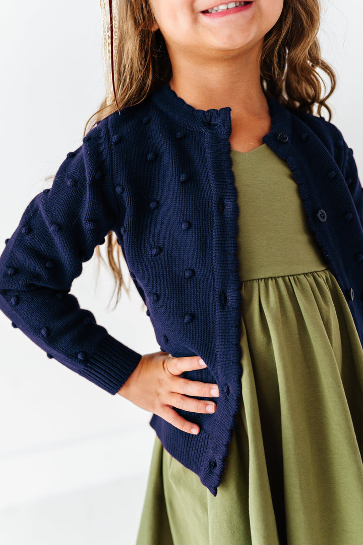 girl wearing navy blue cardigan with 3d raised bubble dots and olive green v-neck dress on white background