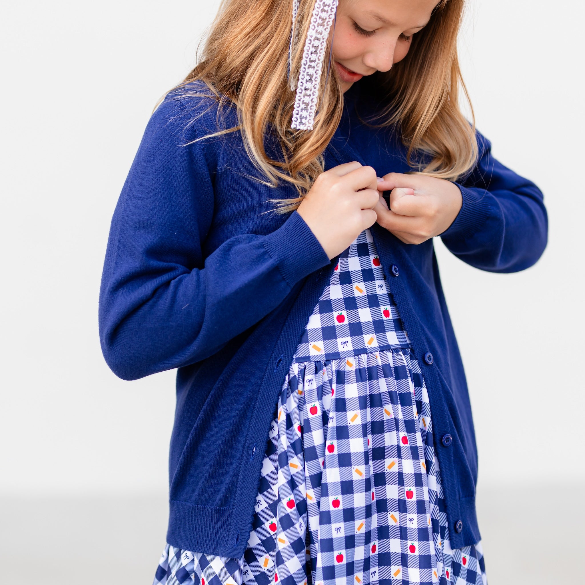 girl buttoning navy cotton cardigan over school themed navy gingham dress with pencils apples and bows on it