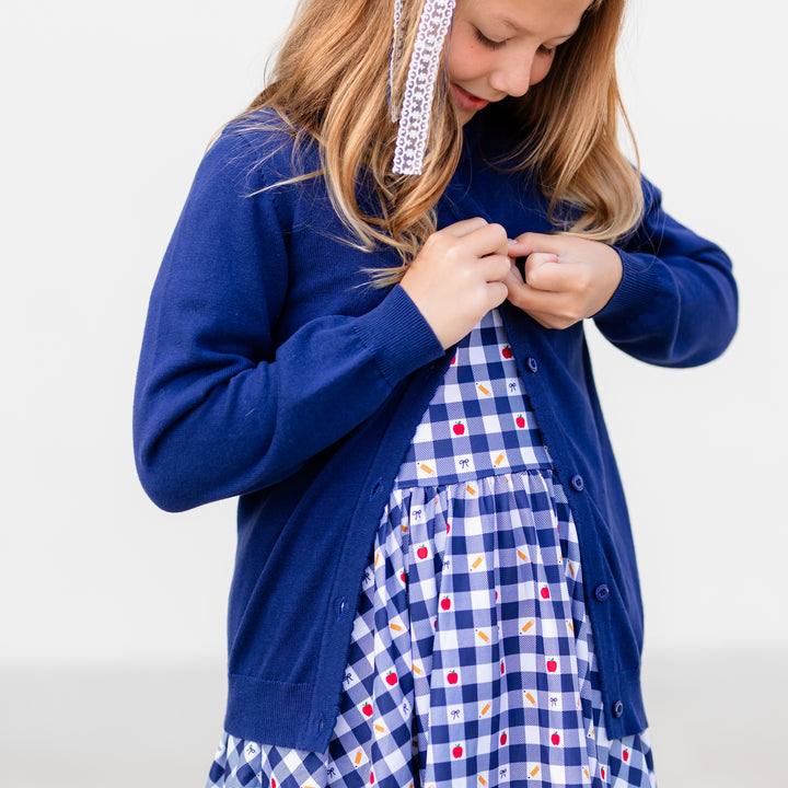 girl buttoning navy cotton cardigan over school themed navy gingham dress with pencils apples and bows on it