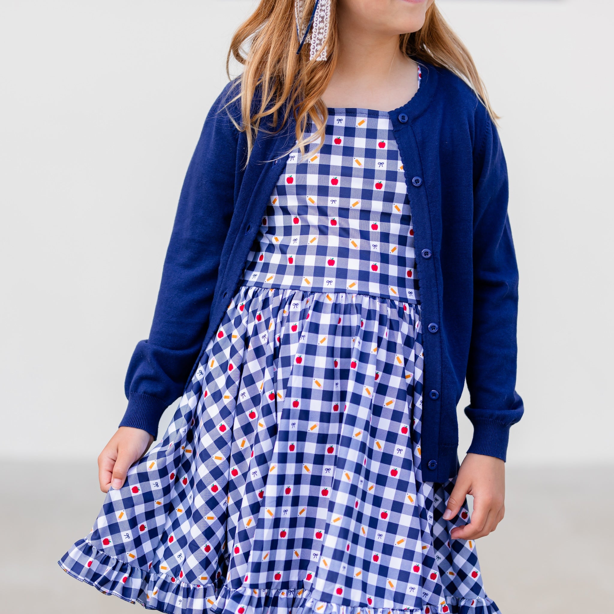 girl in short sleeve, square neck back to school dress in navy and white gingham with apple, pencil and bow print details with navy cardigan sweater