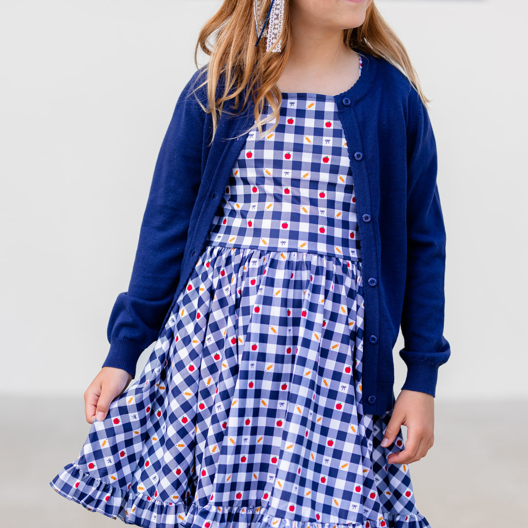 girl in short sleeve, square neck back to school dress in navy and white gingham with apple, pencil and bow print details with navy cardigan sweater