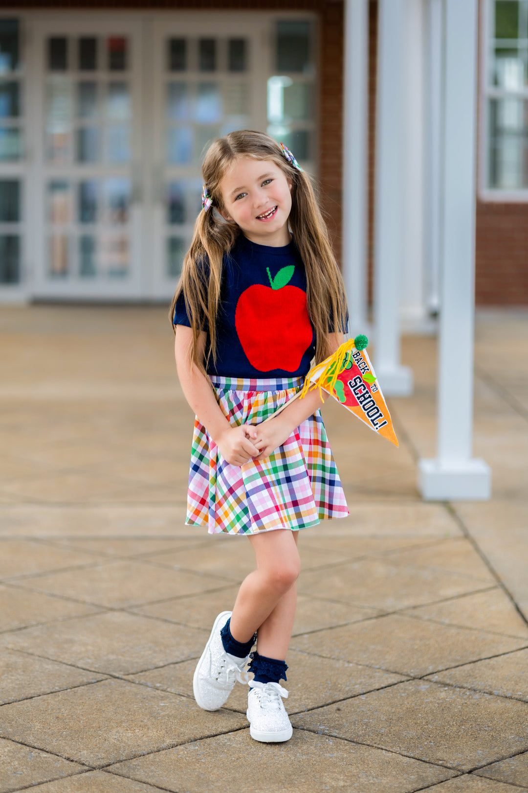 young girl with back-to-school flag wearing navy short sleeve knit apple sweater and coordinating gingham skort in primary colors