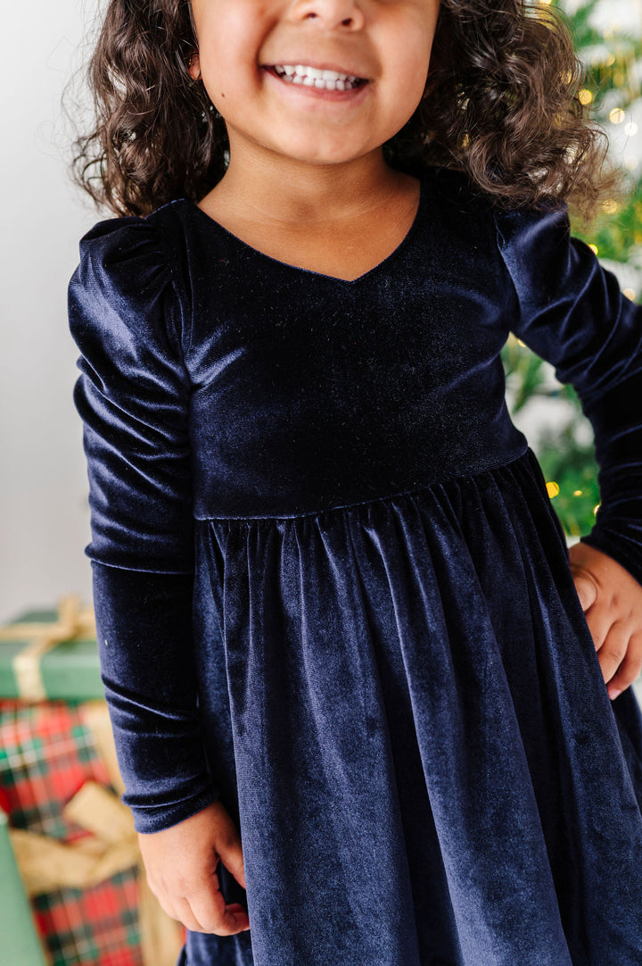 A young girl wearing a long-sleeve navy blue velvet dress, smiling in front of a softly blurred Christmas tree and wrapped gifts.