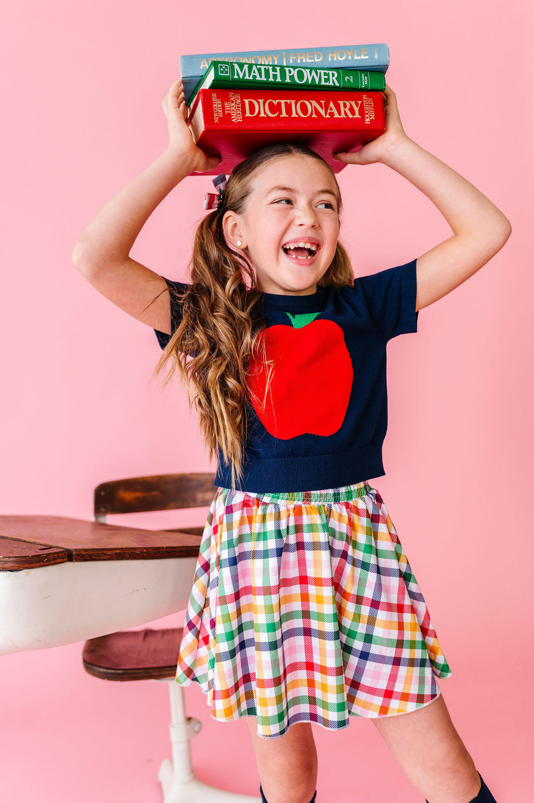 girl wearing rainbow plaid skort with big apple knit top, holding books for back to school