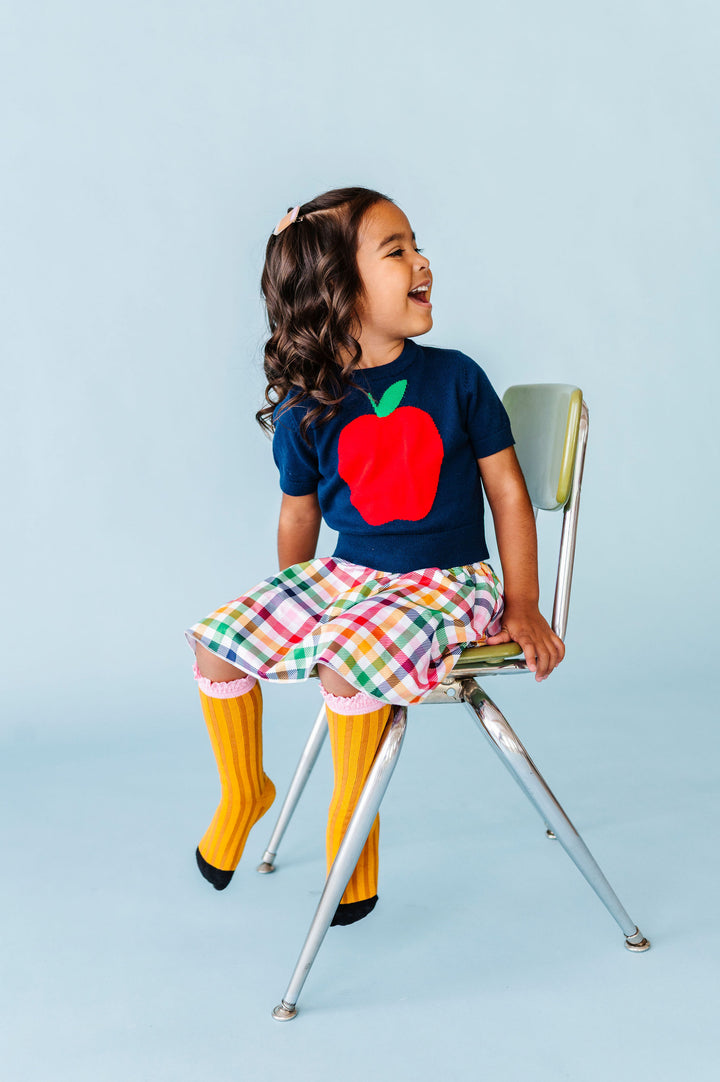 girl wearing back to school outfit with big apple sweater and rainbow gingham skort