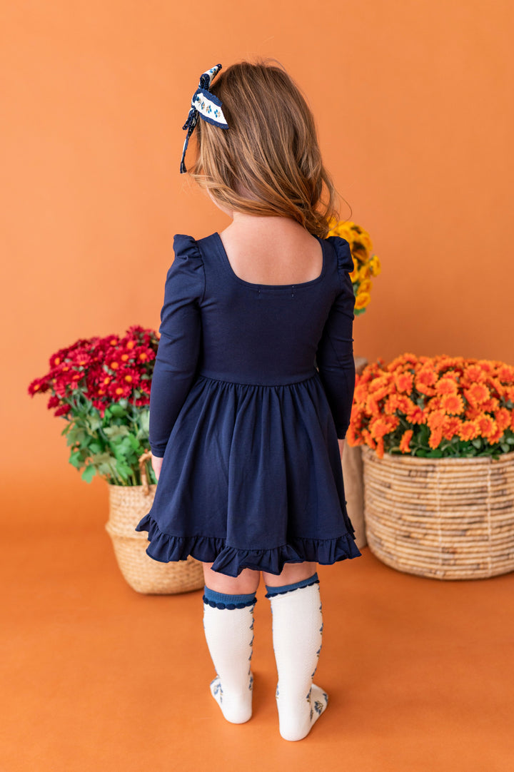 little girl showing back of square neck long sleeve twirl dress in navy paired with matching ivory and blue floral socks and bow