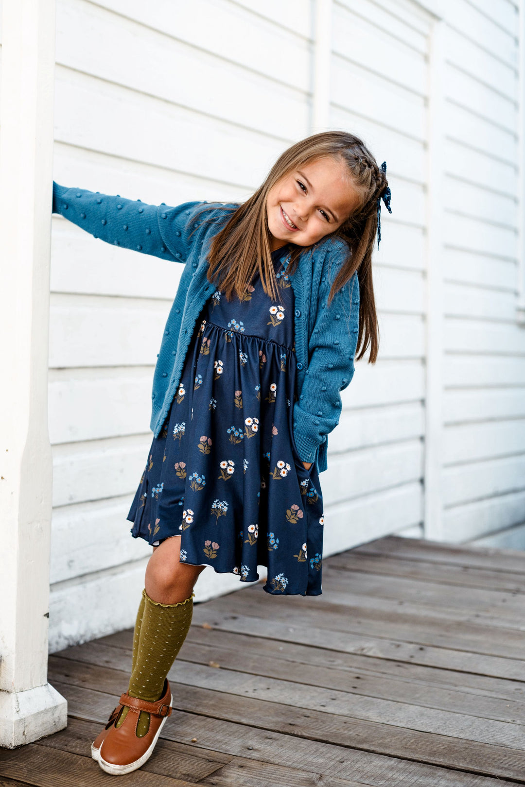 little girl on porch in navy blue floral twirl dress paired with fall blue bubble knit cardigan and olive dotted knee socks with lettuce trim