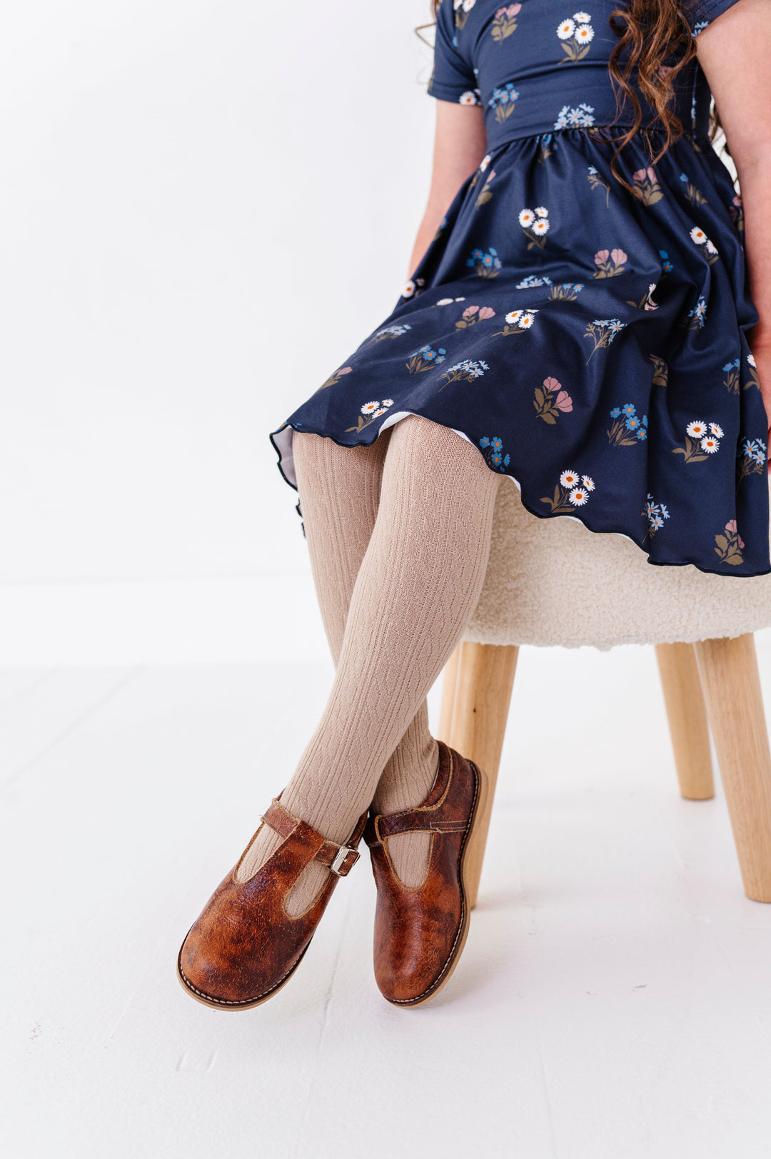 little girl sitting on stool in navy fall floral twirl dress paired with coordinating oat cable knit tights and brown mary janes