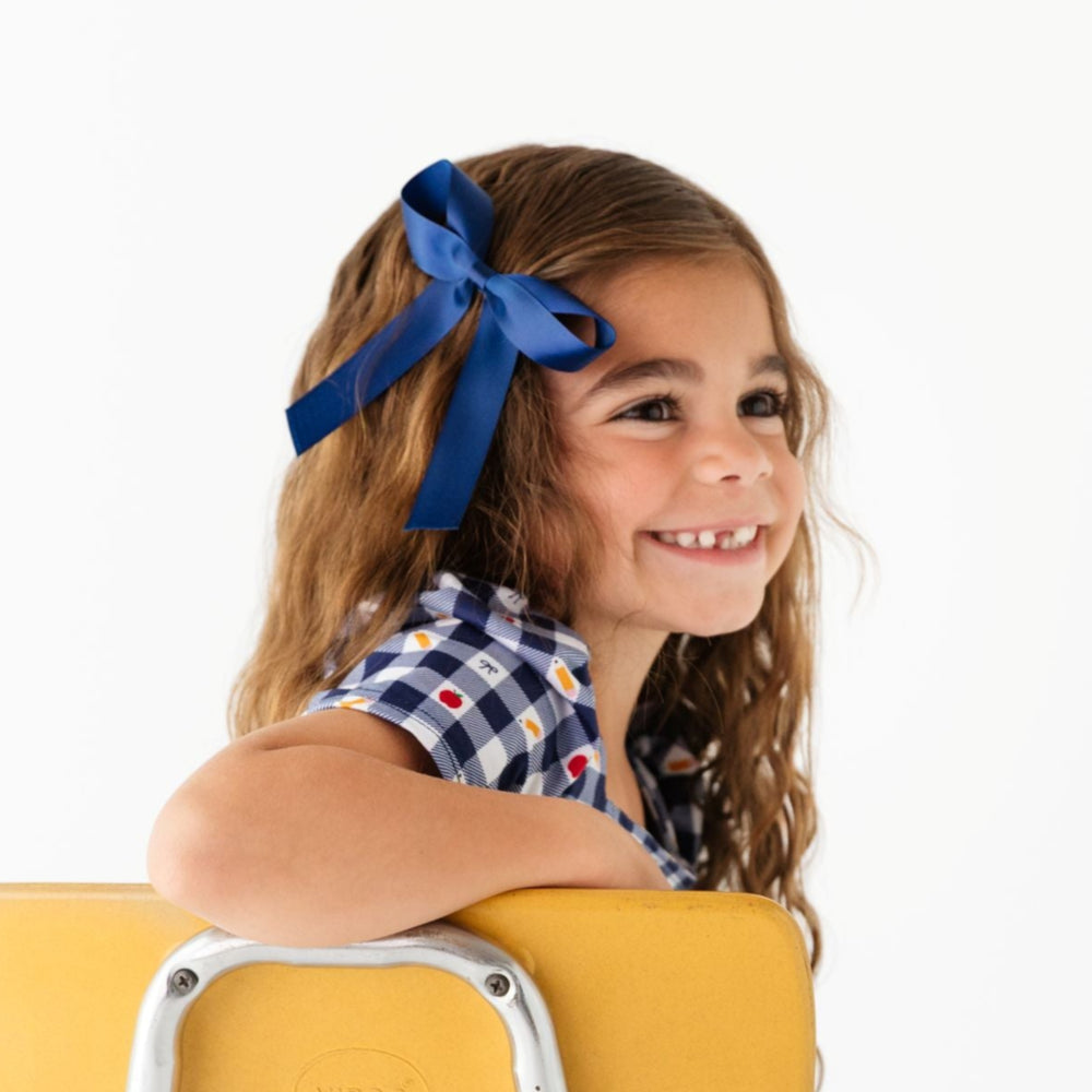 little girl sitting in desk chair with navy blue satin ribbon bow and matching school themed navy gingham twirl dress