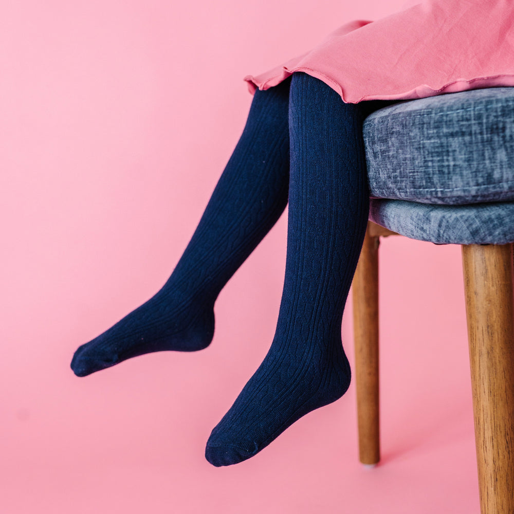 closeup of navy-blue cable knit tights on little girl sitting on chair in pink lettuce trim twirl dress
