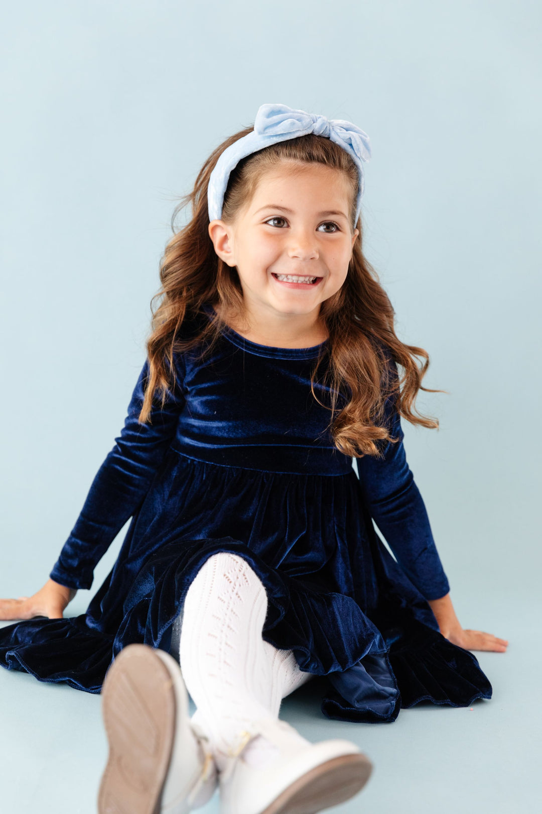 little girl sitting in dark blue long sleeve velvet winter dress, white tights and light blue velvet headband