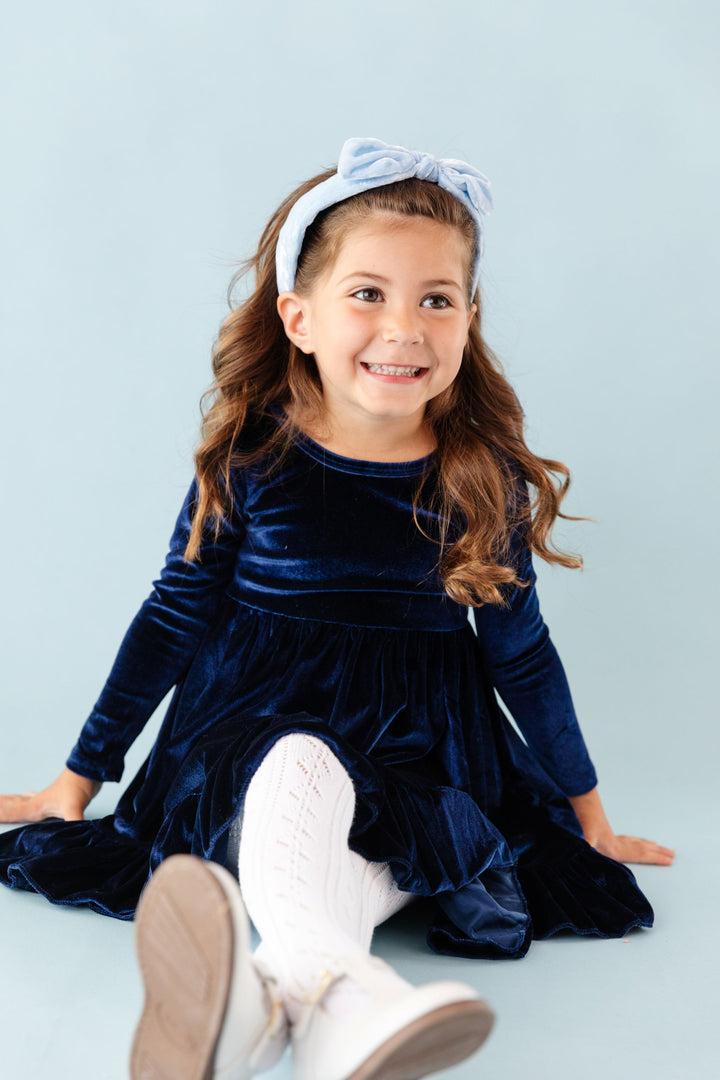 little girl sitting in dark blue long sleeve velvet winter dress, white tights and light blue velvet headband