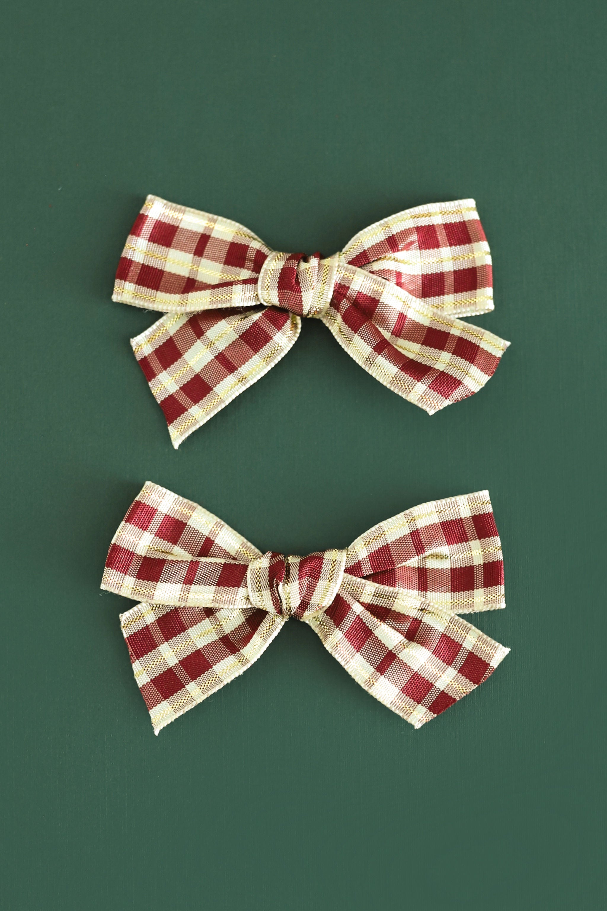 Two matching fabric hair bows with a red and beige gingham plaid pattern, arranged vertically on a solid dark green background.