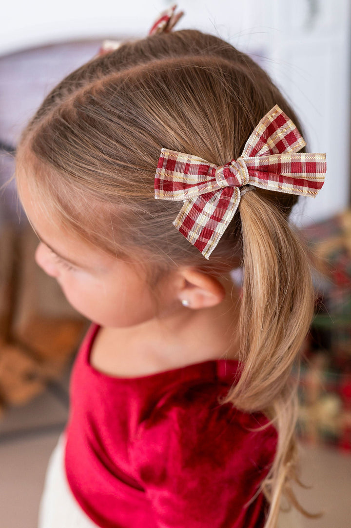 A young girl with light brown hair tied back in a ponytail, wearing a red and white gingham bow hair clip. She is wearing a red velvet top, photographed in a festive indoor setting with a blurred fireplace and holiday decorations in the background.