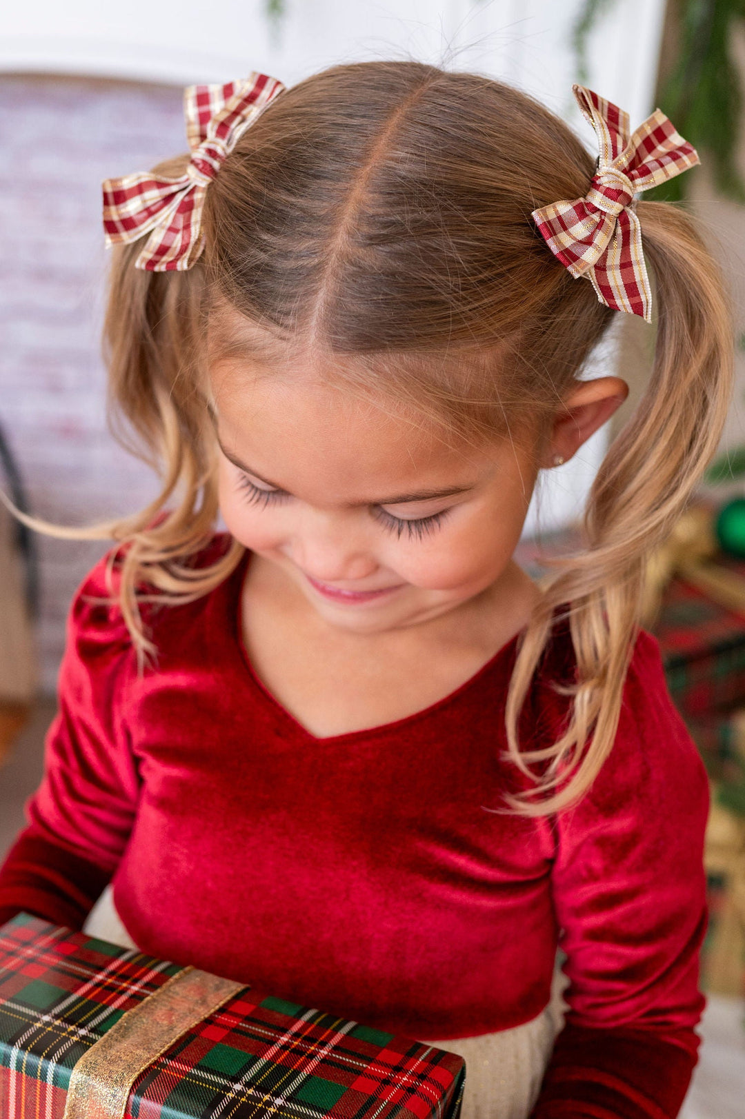 A young girl with blonde pigtails wearing red and cream gingham bows and a red velvet dress, holding a Christmas gift box wrapped in a red, green, and yellow plaid pattern with a gold ribbon. The background shows a cozy indoor holiday setting with a fireplace and a Christmas tree.