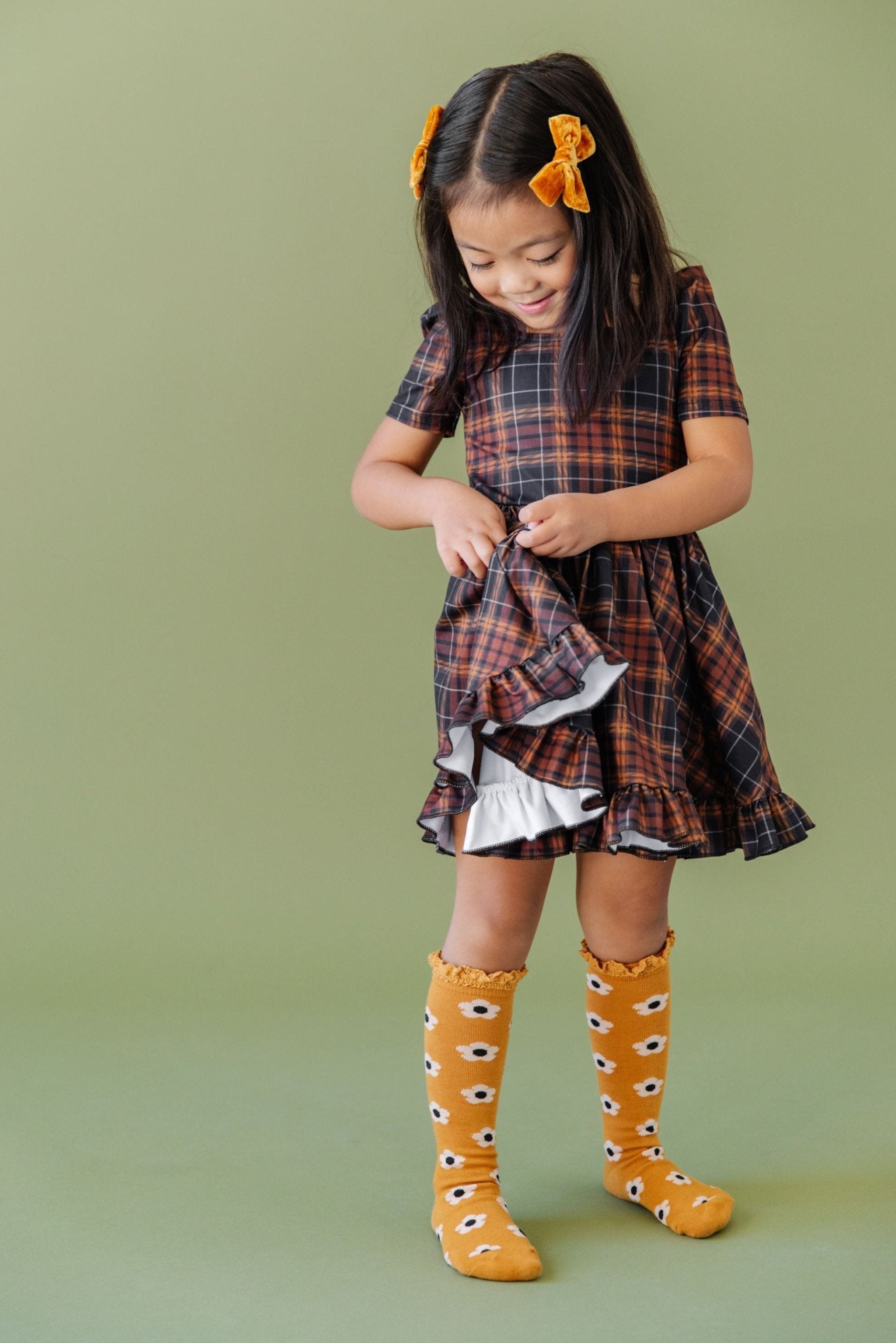 little girl in black and brown fall plaid twirl dress and matching mustard floral knee highs and pigtail bows