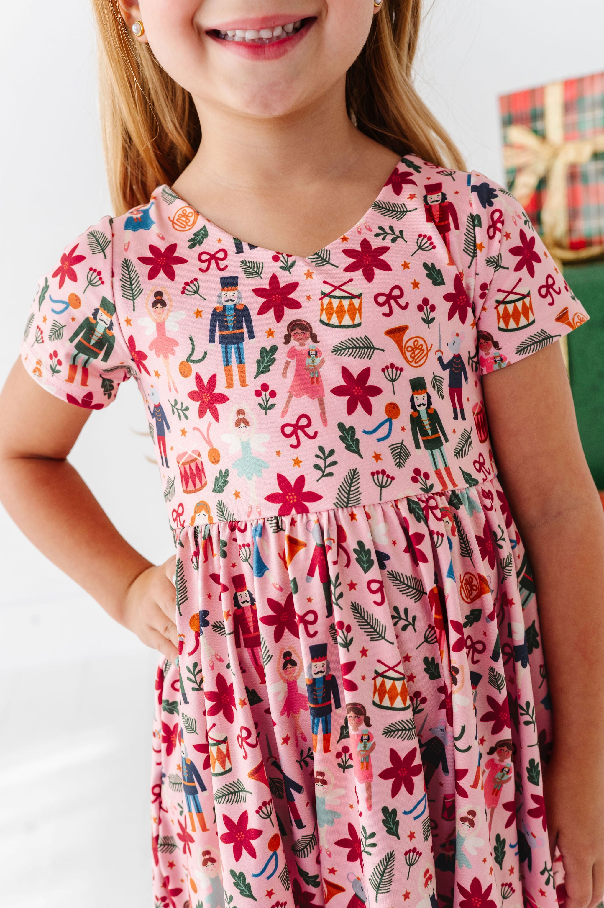A young girl wearing a pink short-sleeve dress with a colorful holiday nutcracker and floral print, shown from the chest down against a bright indoor background.