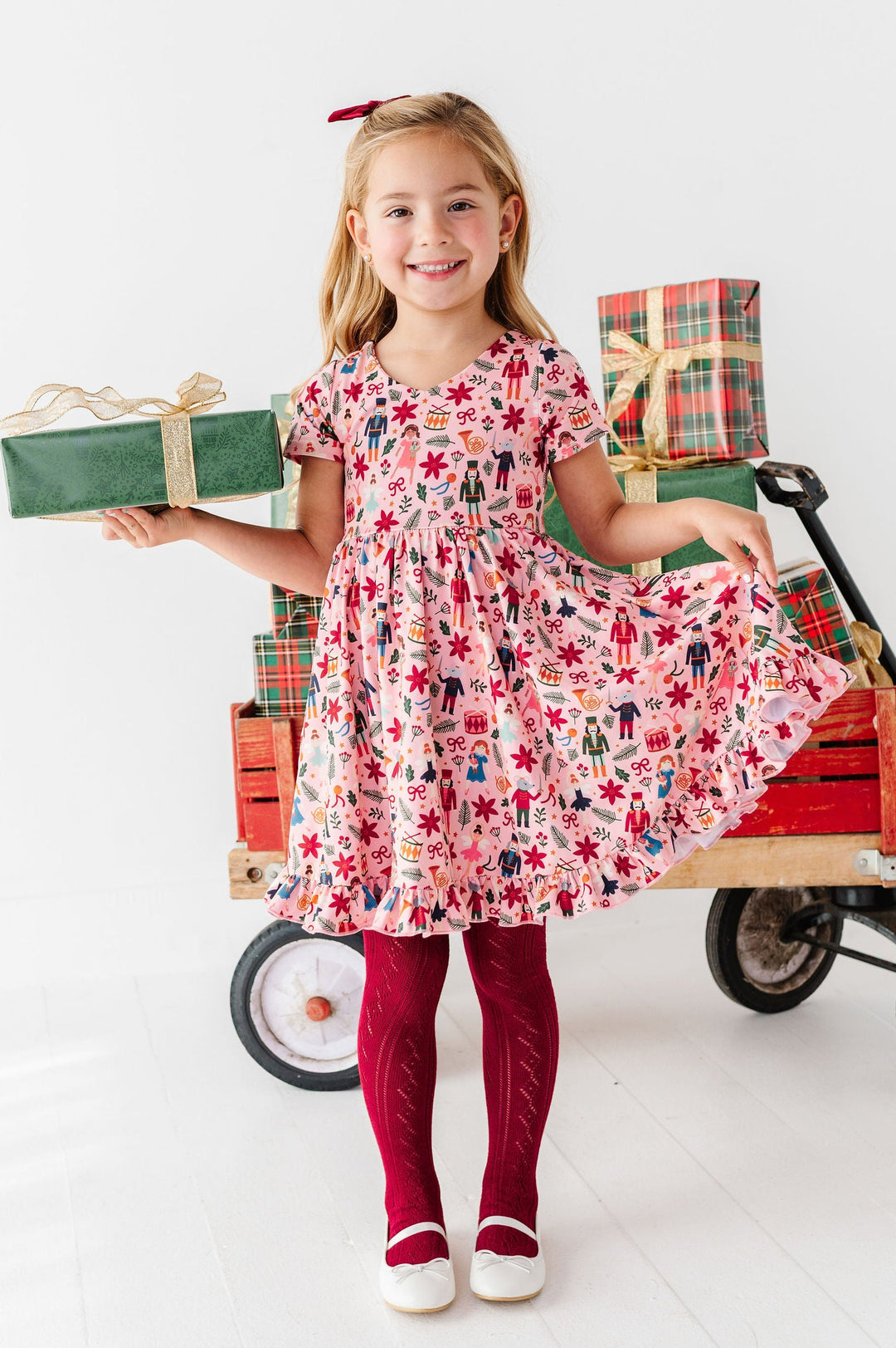 A young girl stands in a bright white studio holding a wrapped gift box next to a vintage-style red wagon loaded with presents. She is wearing a pink holiday print dress, dark red tights, and white shoes.