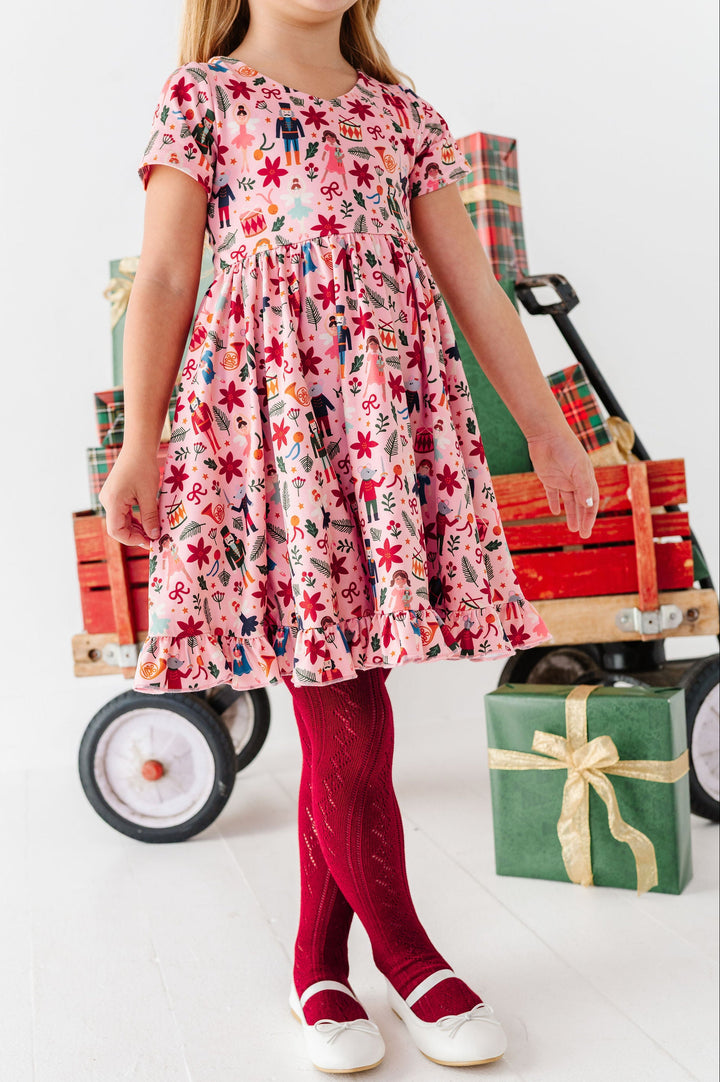 A young girl wearing a pink Christmas-themed dress with a red tights and white shoes, standing next to a wooden wagon filled with wrapped gifts on a white studio floor.