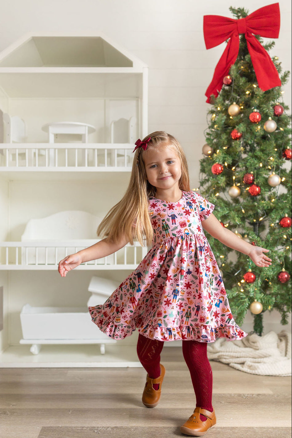 A young girl with long blonde hair wearing a pink floral dress, burgundy tights, and brown shoes is twirling in a brightly lit room next to a decorated Christmas tree and a white dollhouse.