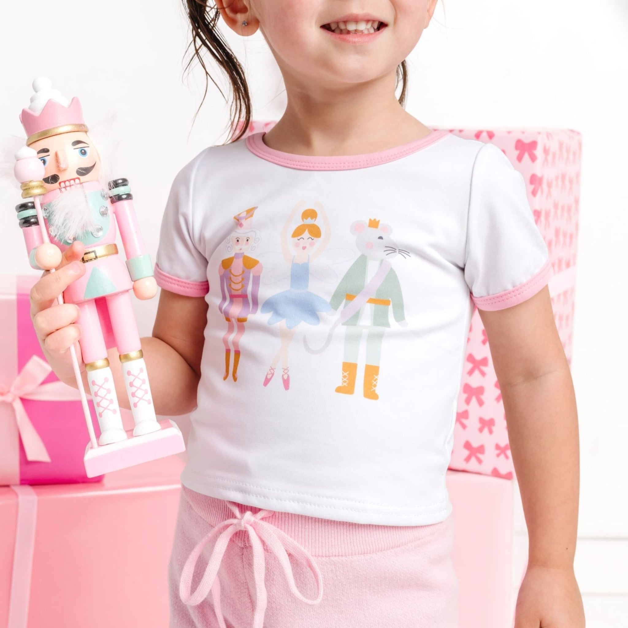 closeup of little girl holding nutcracker toy in white fitted short sleeve top with three nutcracker inspired characters and light pink binding