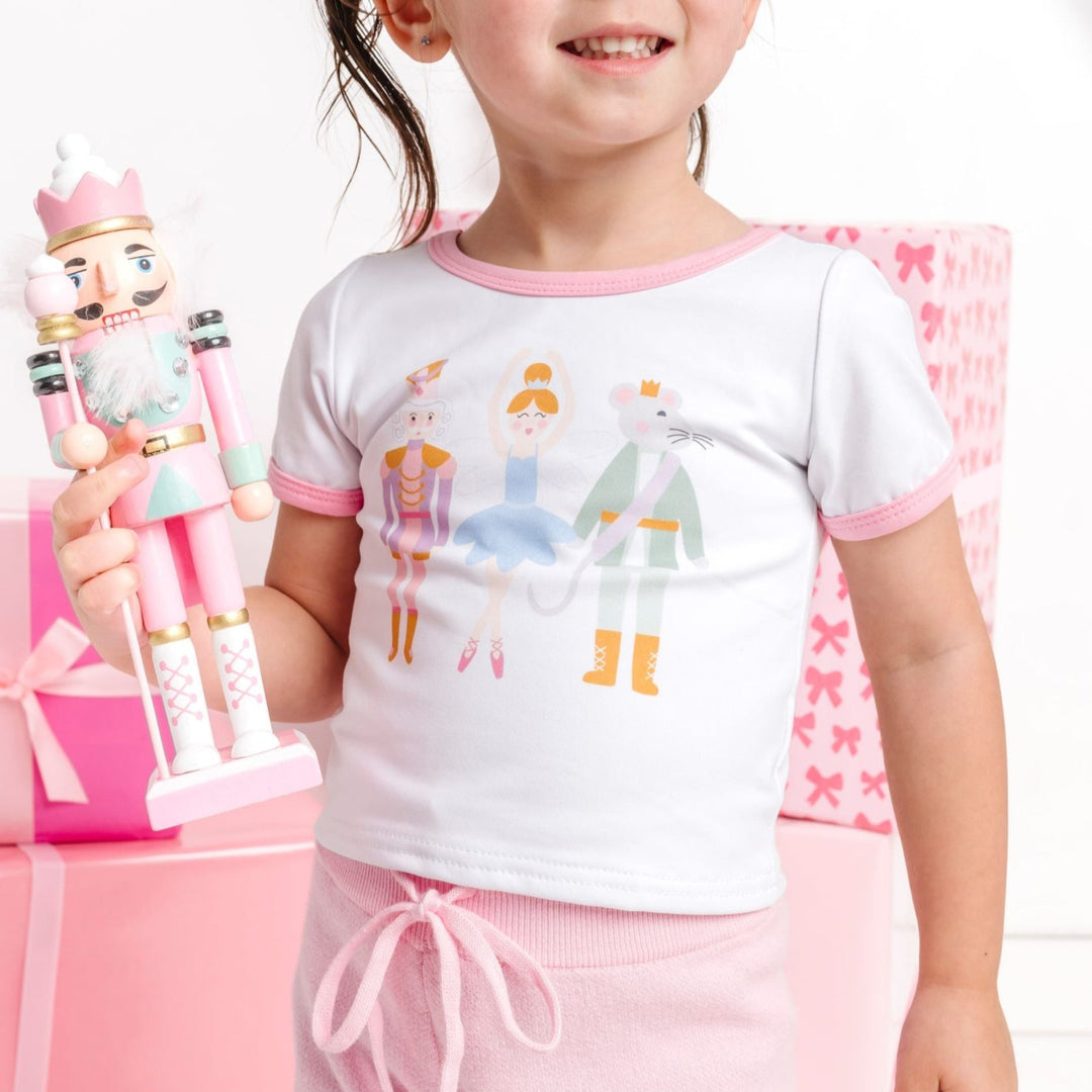 closeup of little girl holding nutcracker toy in white fitted short sleeve top with three nutcracker inspired characters and light pink binding