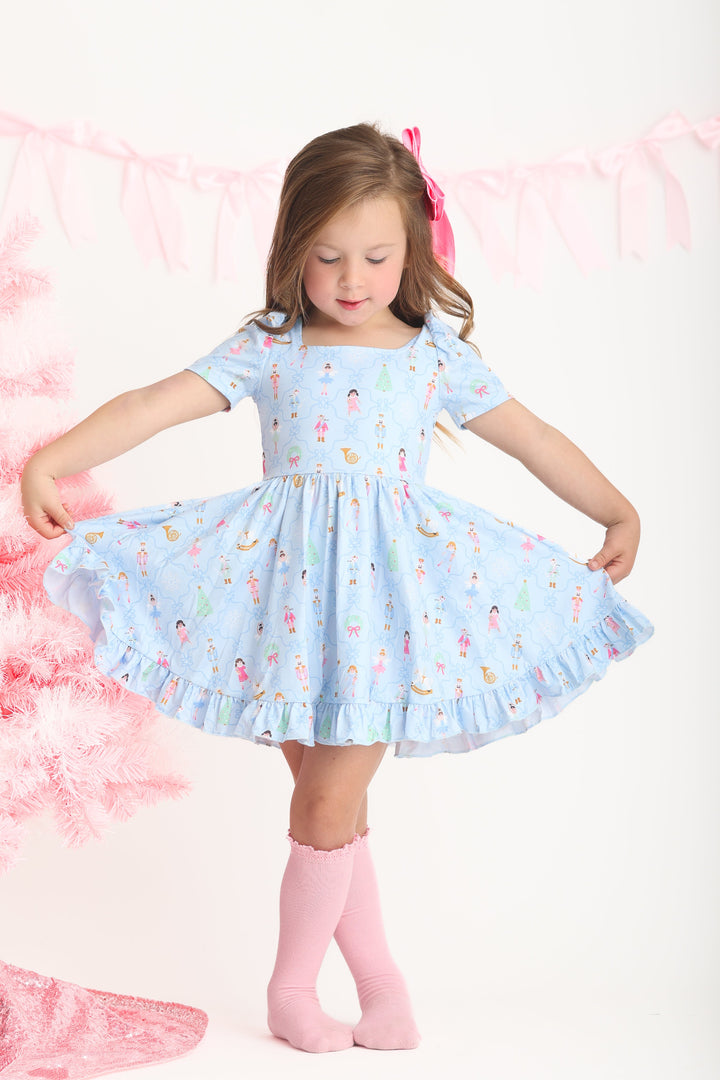 little girl posing by pink Christmas tree in light blue short sleeve twirl dress with Nutcracker inspired print and matching pink satin bow and knee igh socks