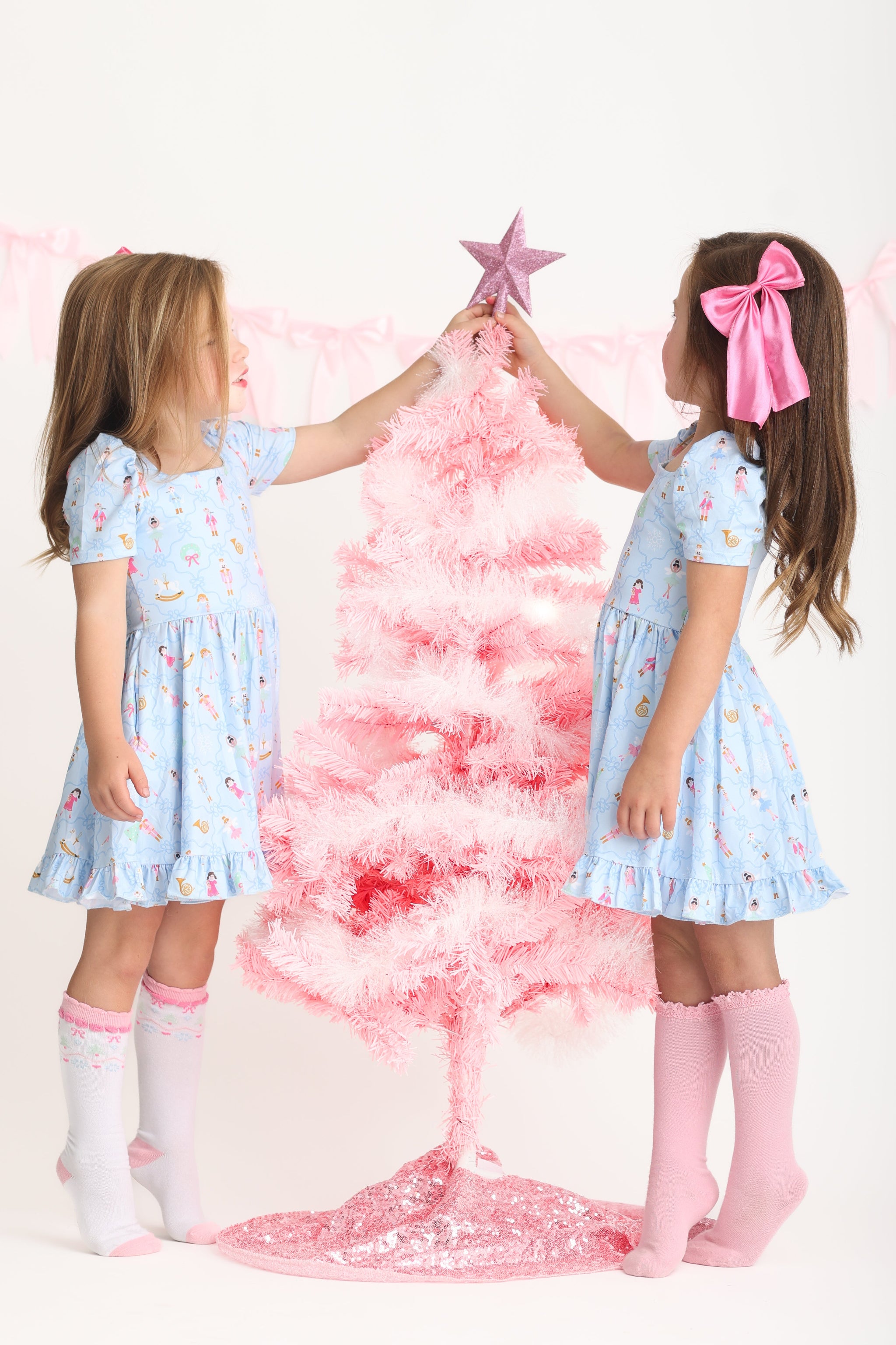 sisters decorating pink Christmas tree in blue nutcracker print twirl dress and matching satin bow and socks 
