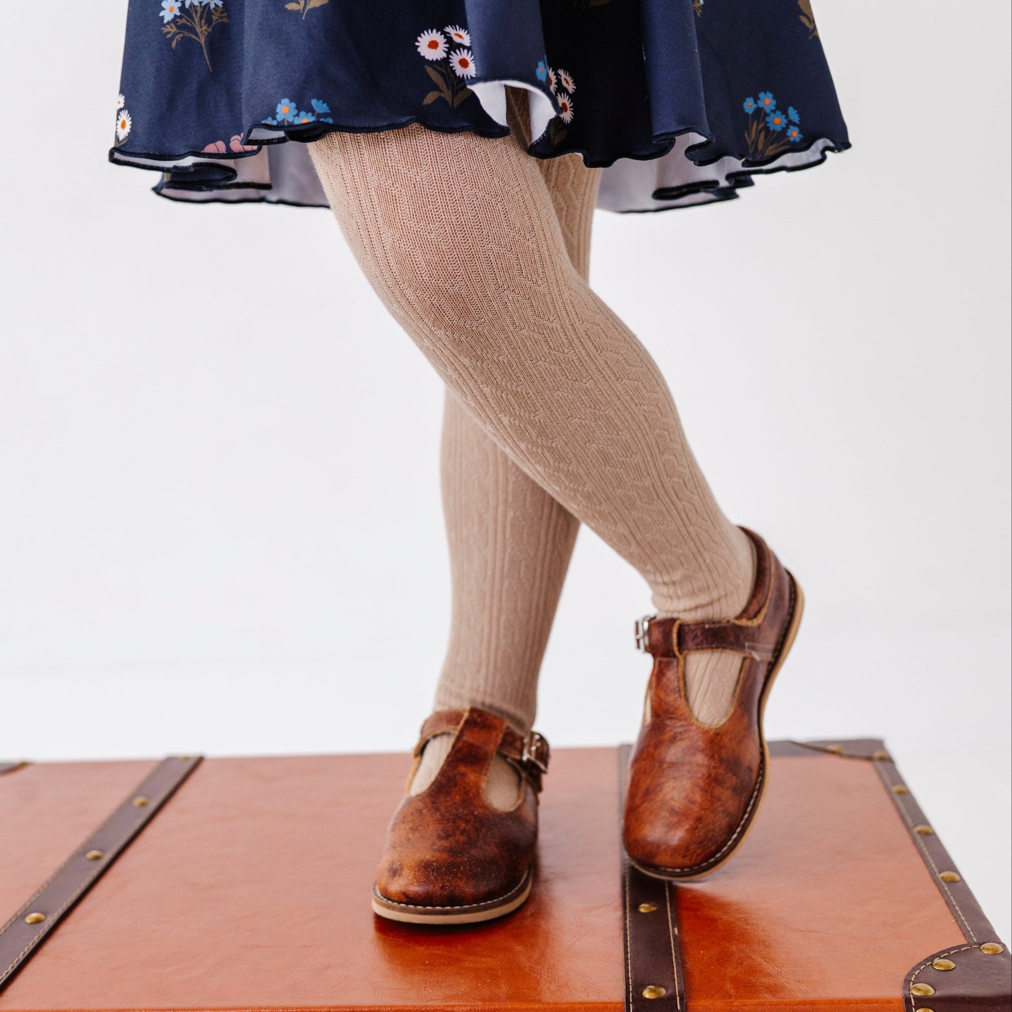 little girl standing on vintage suitcase in oat cable knit tights paired with navy floral twirl dress and brown mary janes