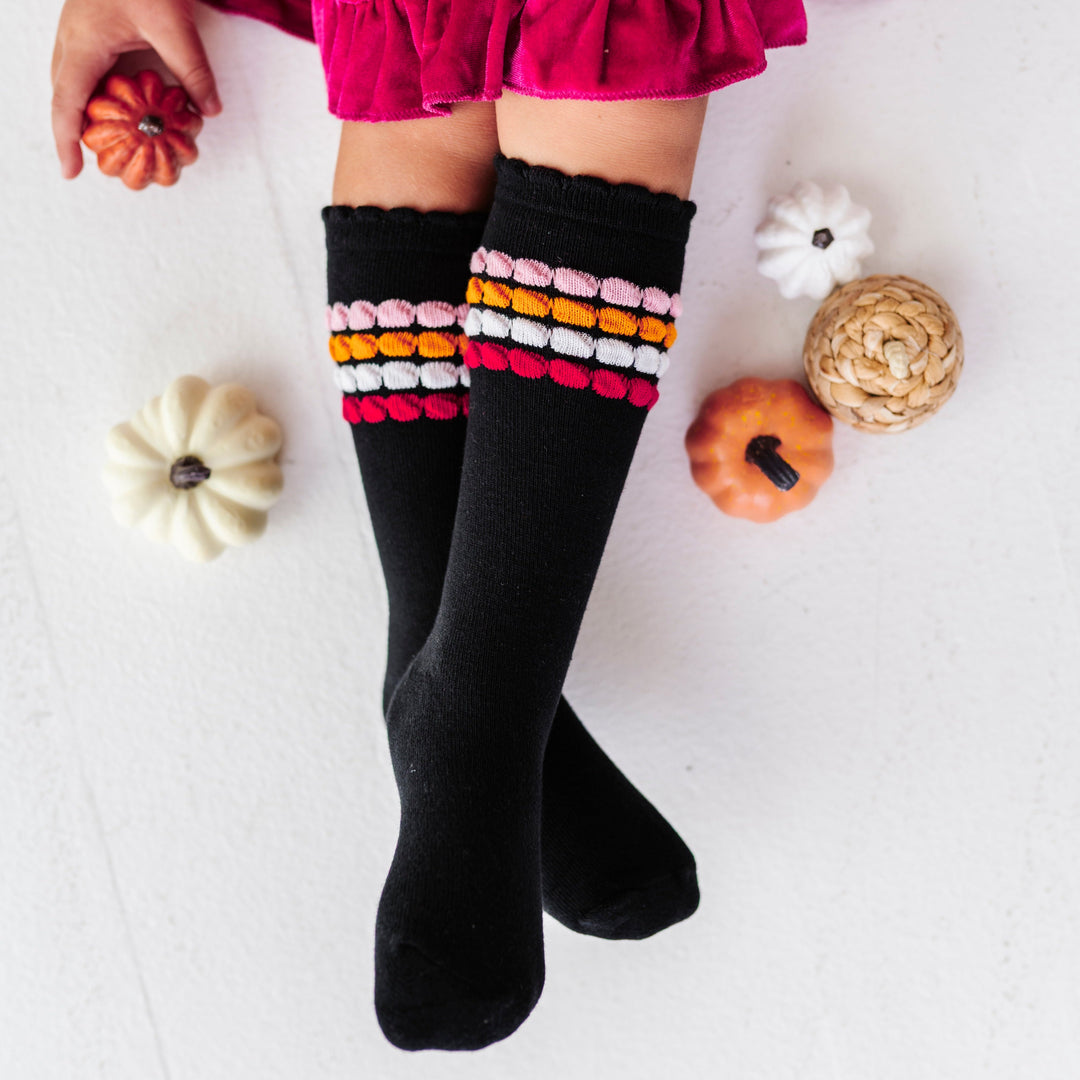 young girl holding pumpkin decor in hot pink velvet twirl dress paired with black knee socks with pink and orange bubble stripes at the calf and lace trim