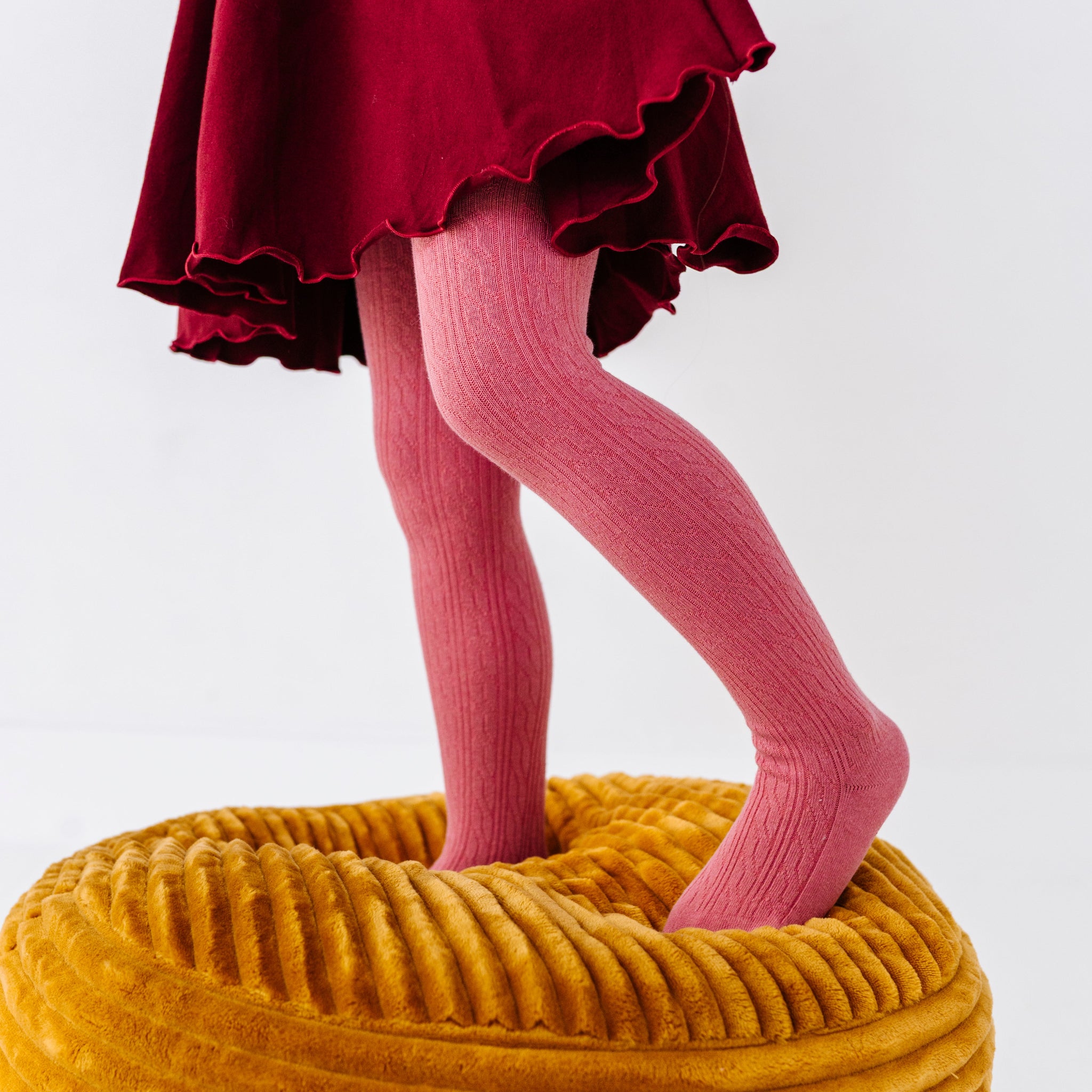 little girl standing on a mustard yellow cushion in fall pink cable knit tights paired with burgundy twirl dress