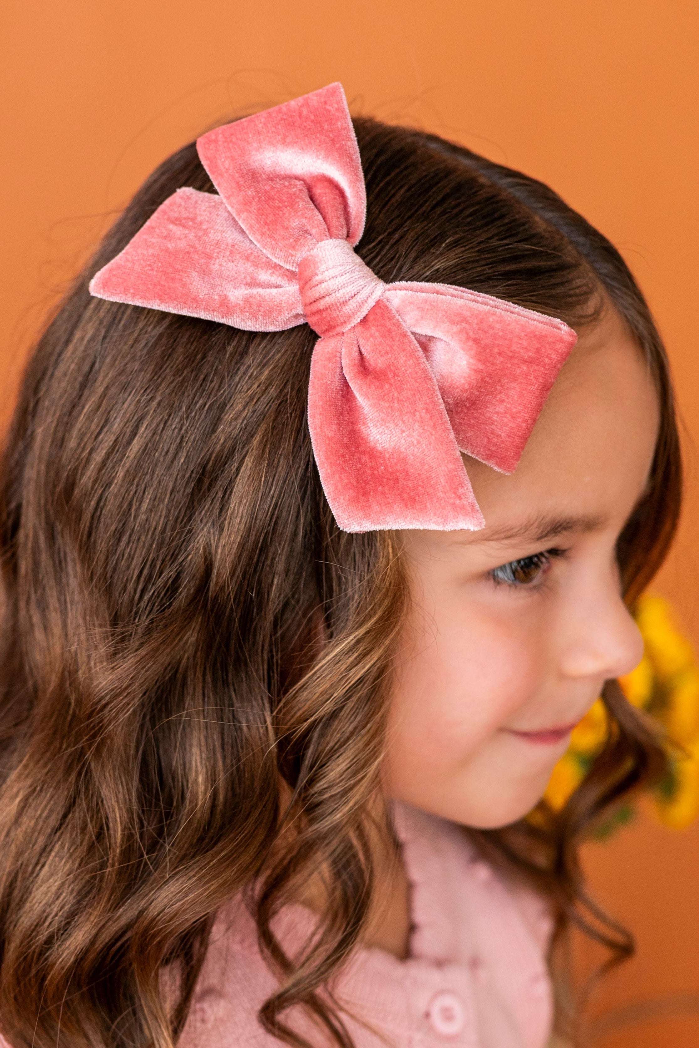 young girl in front of orange backdrop in pink velvet hair bow paired with light pink fall cardigan