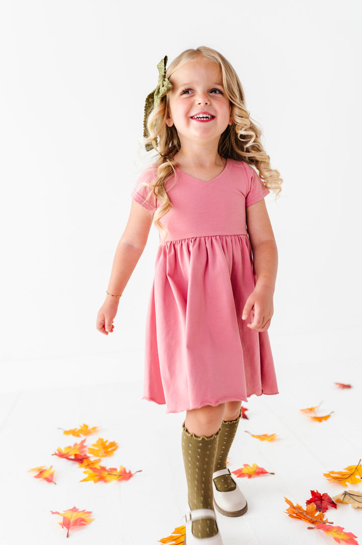 little girl walking in fall leaves in bright mauve v-neck twirl dress paired with olive floral hair bow and dotted knee socks 