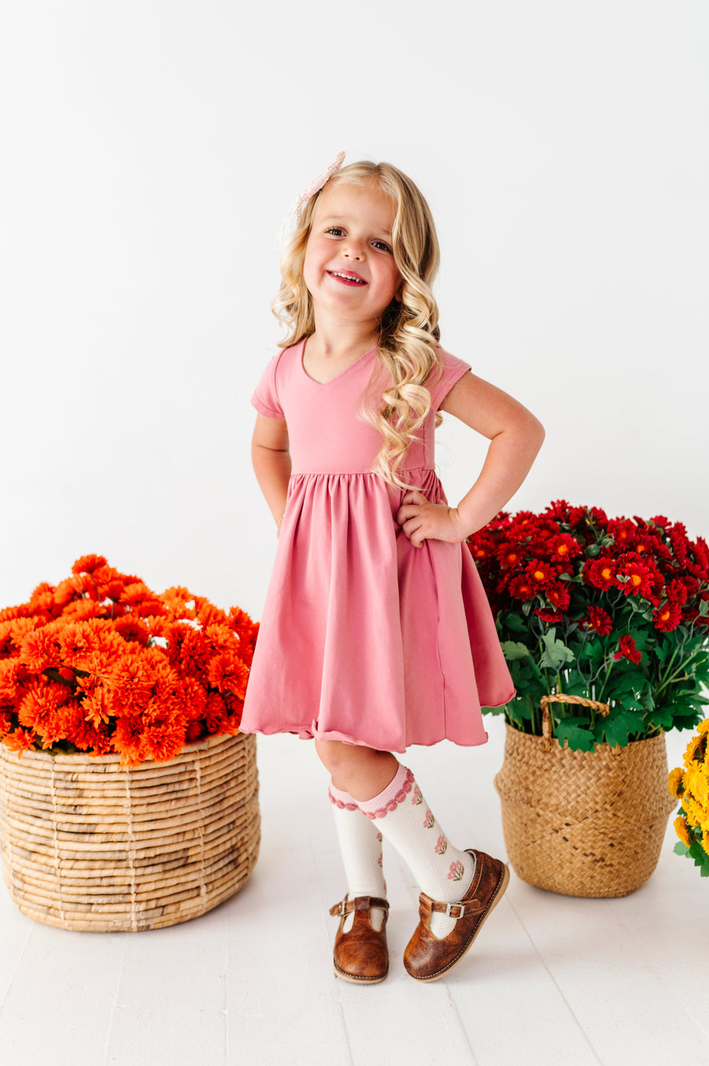 young girl by fall flowers in baskets in rosy, mauve cotton blend twirl dress paired with matching floral knee socks and brown mary jane shoes