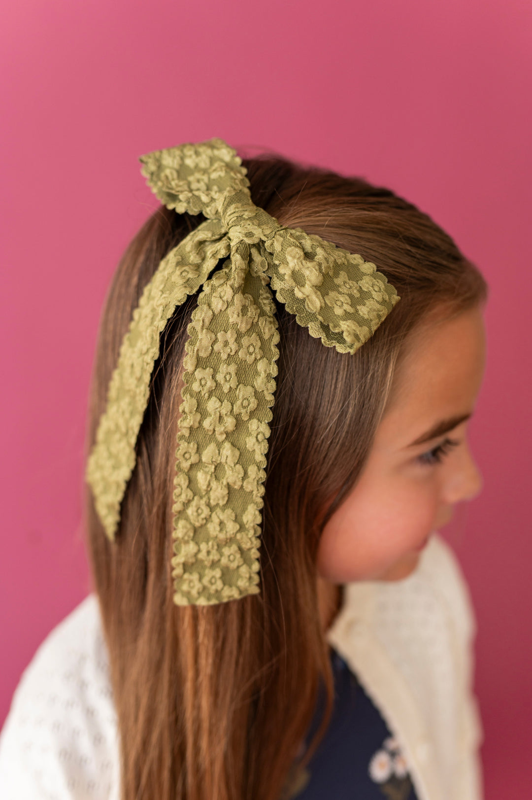 young girl in olive green hair bow with flower design and scalloped edges paired with navy floral dress and ivory cardigan