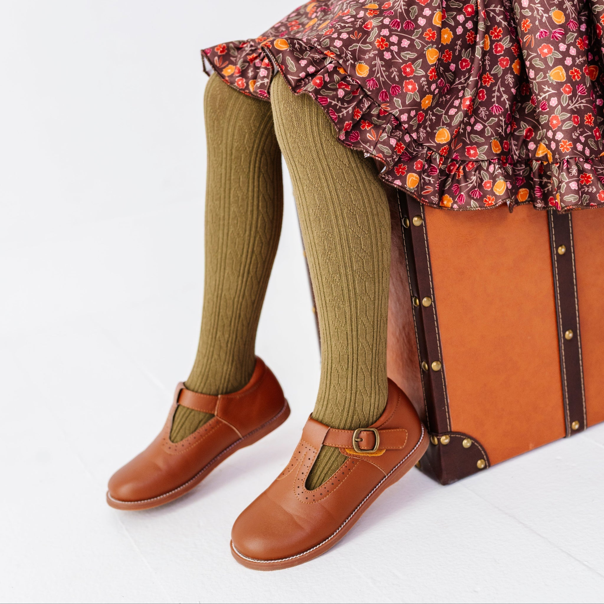 young girl sitting on vintage suitcase in brown fall floral twirl dress paired with olive cable knit tights and brown mary jane shoes