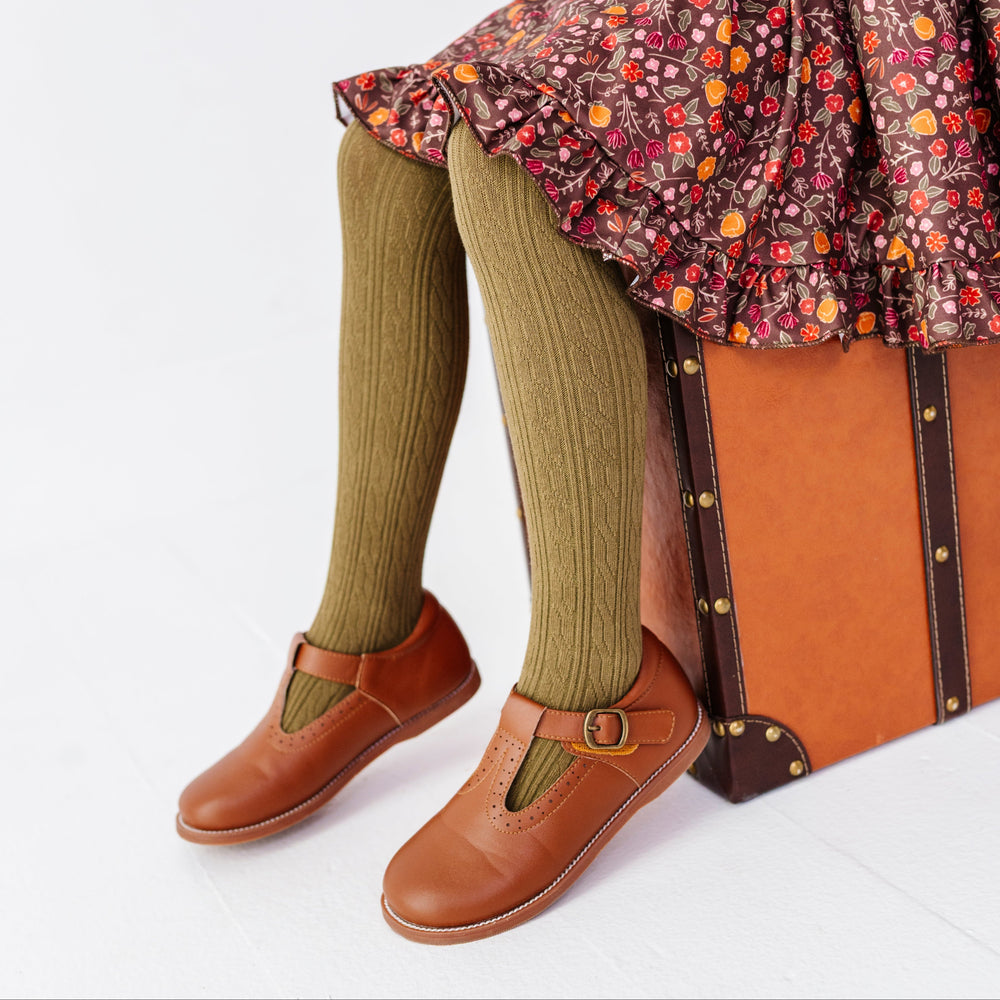 young girl sitting on vintage suitcase in brown fall floral twirl dress paired with olive cable knit tights and brown mary jane shoes