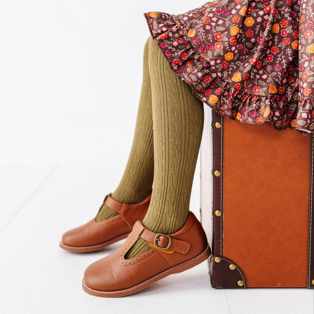 young girl sitting on vintage suitcase in brown fall floral twirl dress paired with olive cable knit tights and brown mary jane shoes