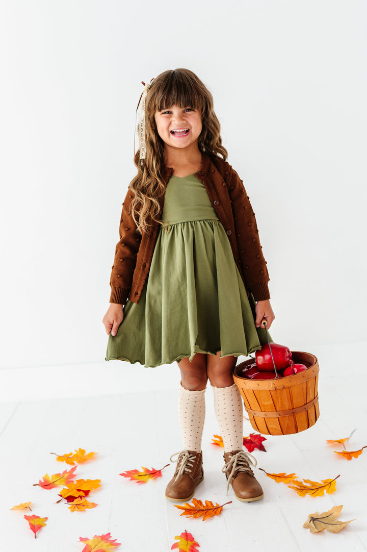 young girl holding basket of apples in olve green twirl dress, brown cardigan with bubble embellishments, cream dotted knee socks and matching lace ribbon bow