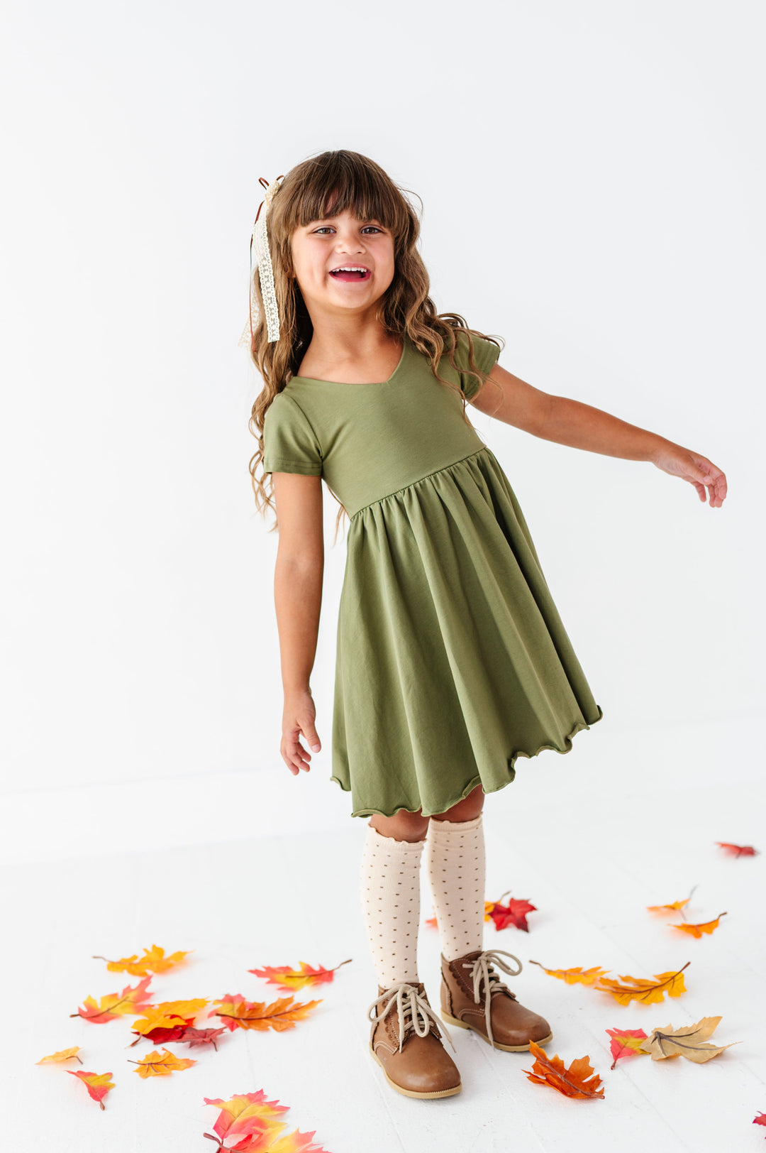young girl standing in fall leaves in solid olive v-neck twirl dress paired with cream with brown dot knee socks and matching cream and brown lace ribbon hair bow