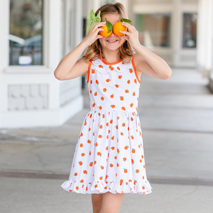 little girl holding two oranges up to her eyes wearing cute orange print tank top summer dress