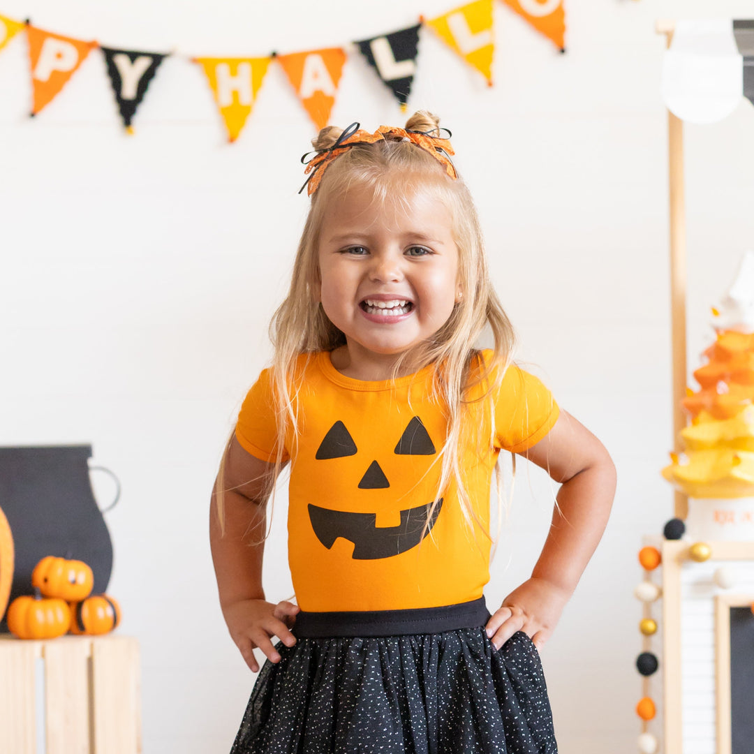 little girl in Halloween-themed outfit with pumpkin shirt and black skirt, surrounded by decorations.