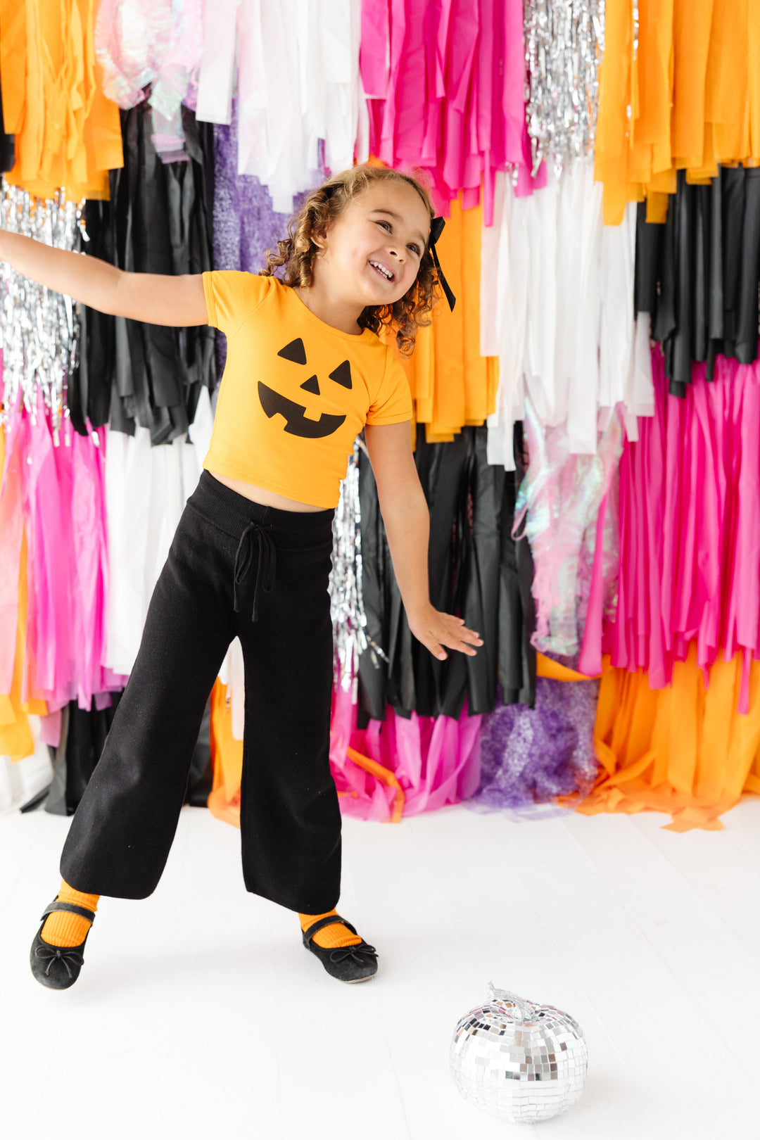 little girl in orange fitted pumpkin face top, black cotton knit sweater pants, orange midi socks and black mary janes