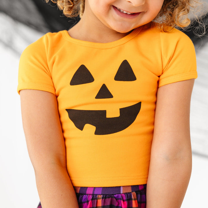 little girl in orange fitted top with black jack-o-lantern face design and Halloween plaid print skort
