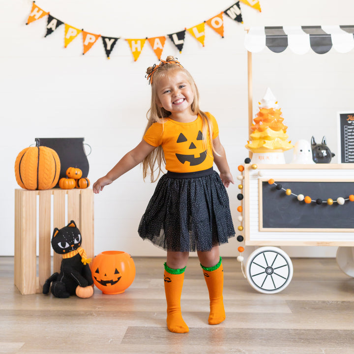 little girl surrounded by Halloween decor wearing orange tee with black Jack-o-lantern face, black sparkle tutu skort, matching orange pumpkin knee high socks and lace bows