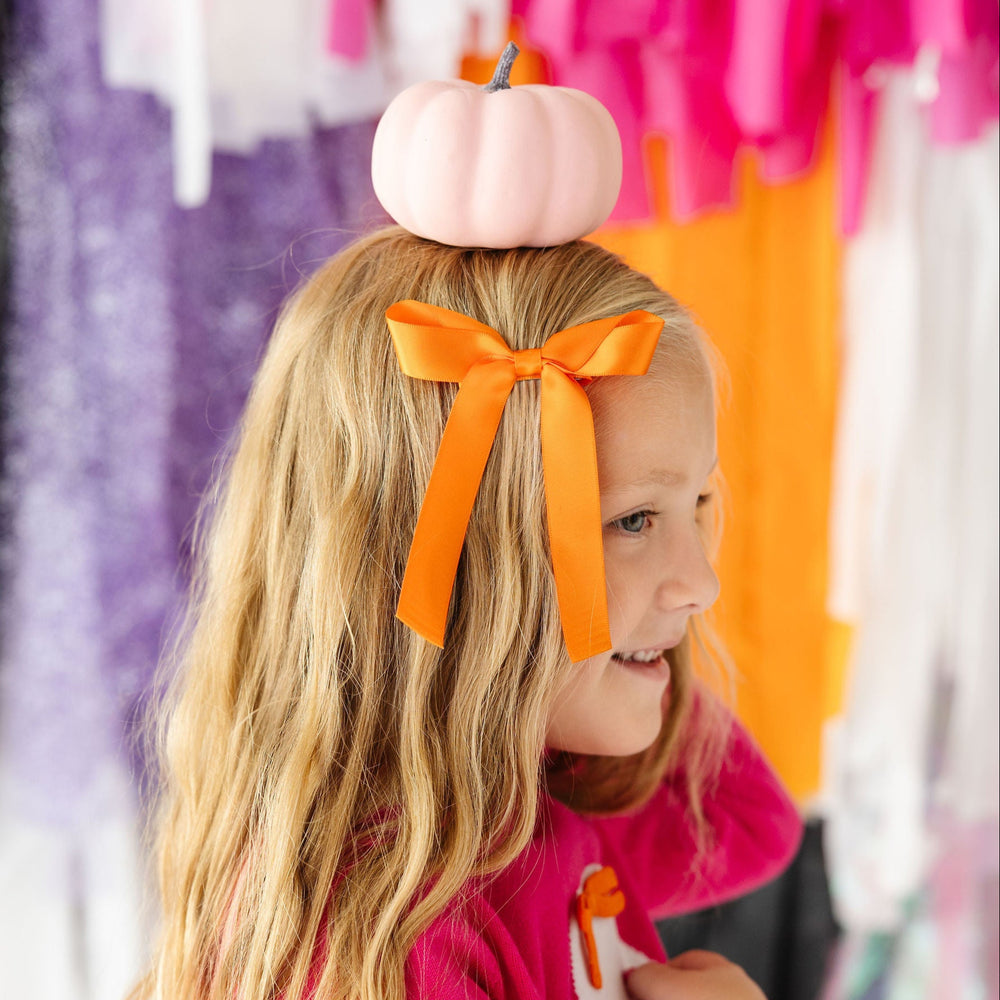 Young girl with a pumpkin on her head and an orange bow in her hair, standing in front of colorful clothing racks.