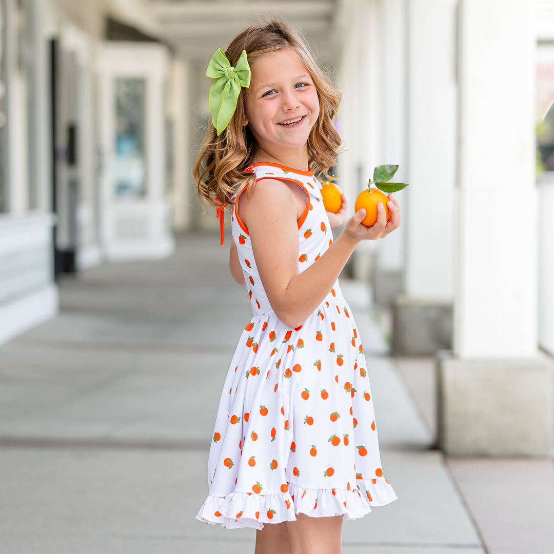 side view of little girl wearing orange print dress with bright green bow in her hair and holding up an orange in each hand
