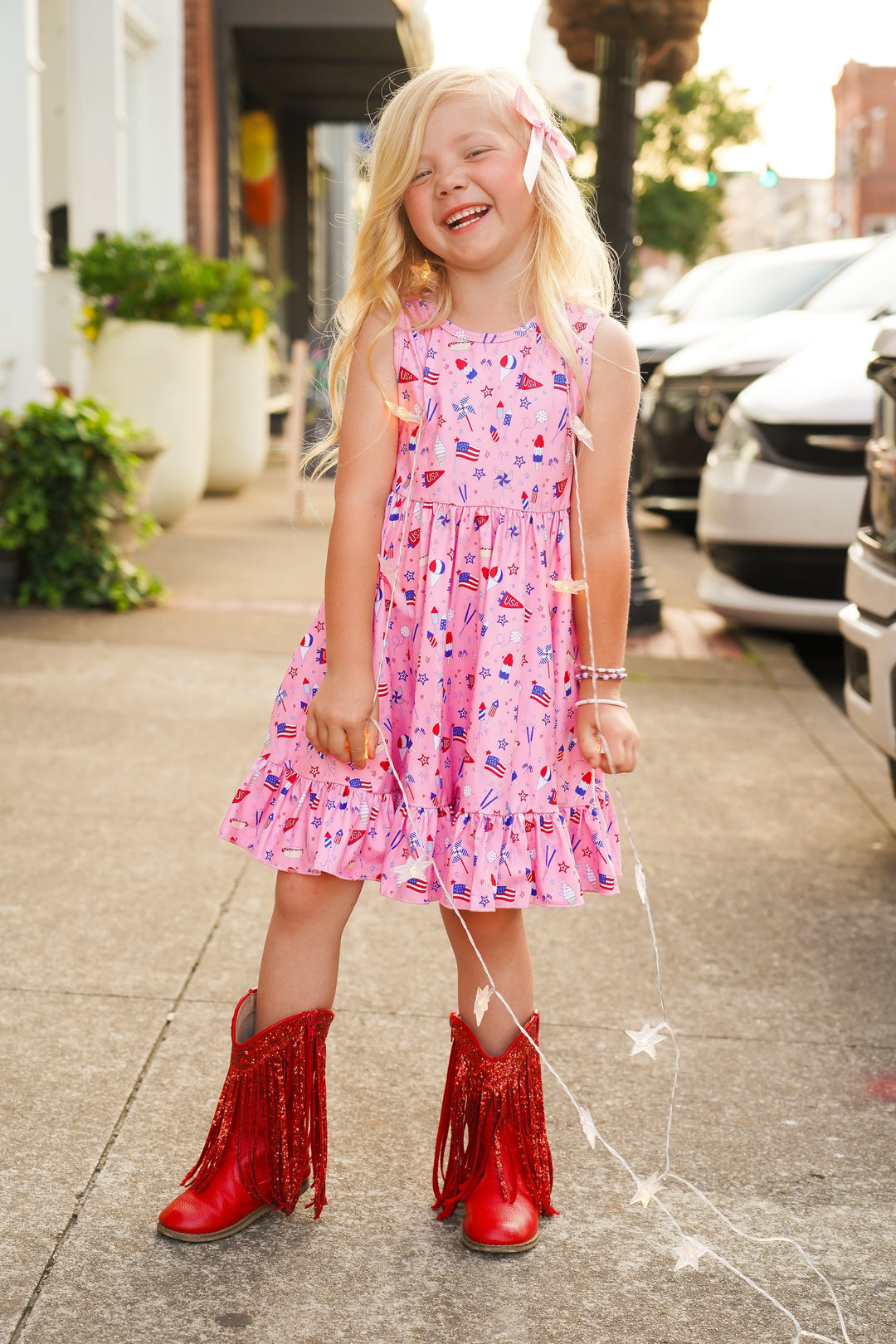 girl wearing pink dress with red white and blue fourth of july print on it and red cowboy boots and matching pink bow