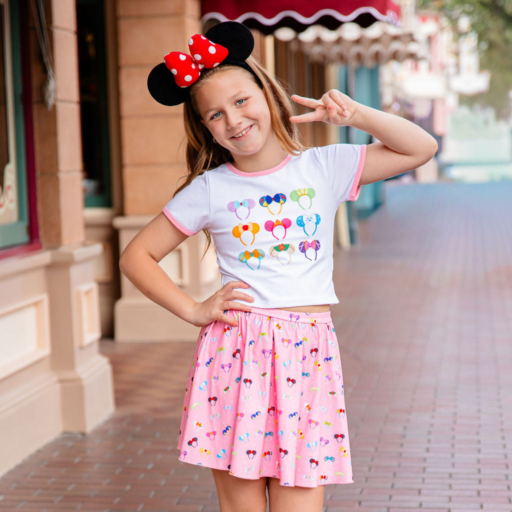Young girl wearing a colorful shirt and pink skirt with cartoon character design, standing on a brick sidewalk.