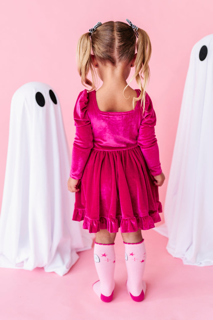 little girl beside ghost decorations facing away from camera to show square neck on the back of her long sleeve, hot pink velvet twirl dress paired with pink ghost knee socks
